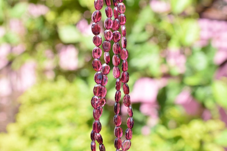 May include: A strand of deep red gemstone beads, likely garnet, strung on a clear thread. The beads are oval-shaped and have a slightly faceted appearance. The background is blurred and out of focus, suggesting a natural setting.