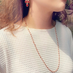 May include: A gold chain necklace with small red beads. The necklace is worn by a person with long brown hair. The person is also wearing a pair of dangling earrings with red gemstones.
