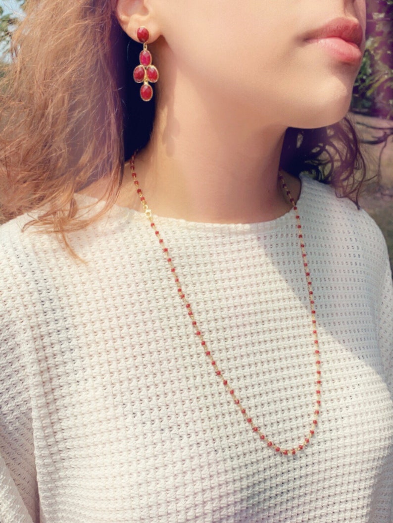 May include: A gold chain necklace with small red beads. The necklace is worn by a person with long brown hair. The person is also wearing a pair of dangling earrings with red gemstones.