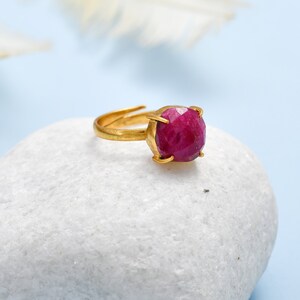 May include: A gold ring featuring a large, faceted ruby gemstone. The ring is displayed on a white stone.