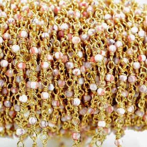 May include: A close-up of a gold chain with pink and white beads. The chain is made of small, delicate links and the beads are faceted and have a marbled appearance.