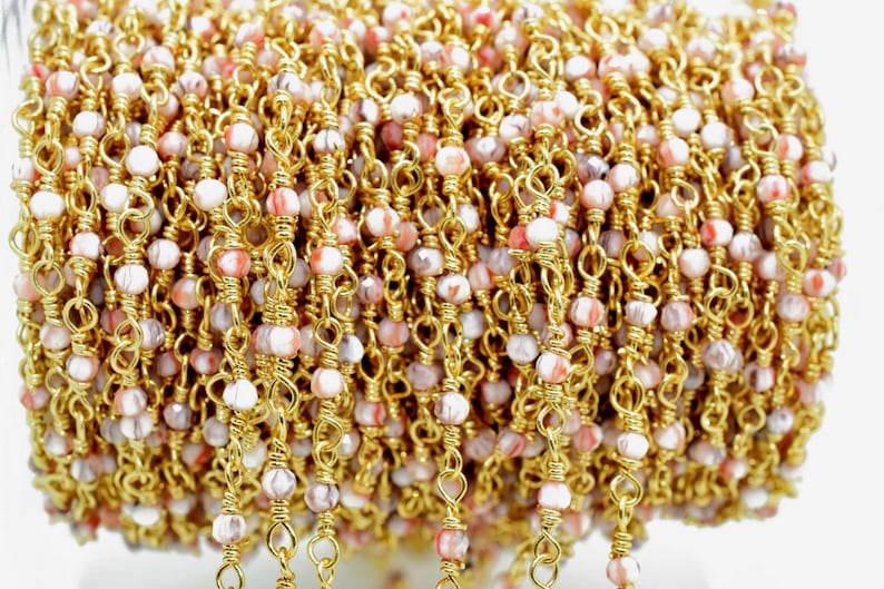 May include: A close-up of a gold chain with pink and white beads. The chain is made of small, delicate links and the beads are faceted and have a marbled appearance.