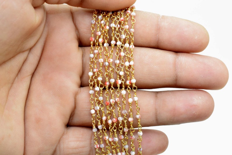 May include: A gold chain with small white and pink beads. The chain is laid out in a straight line on a hand.