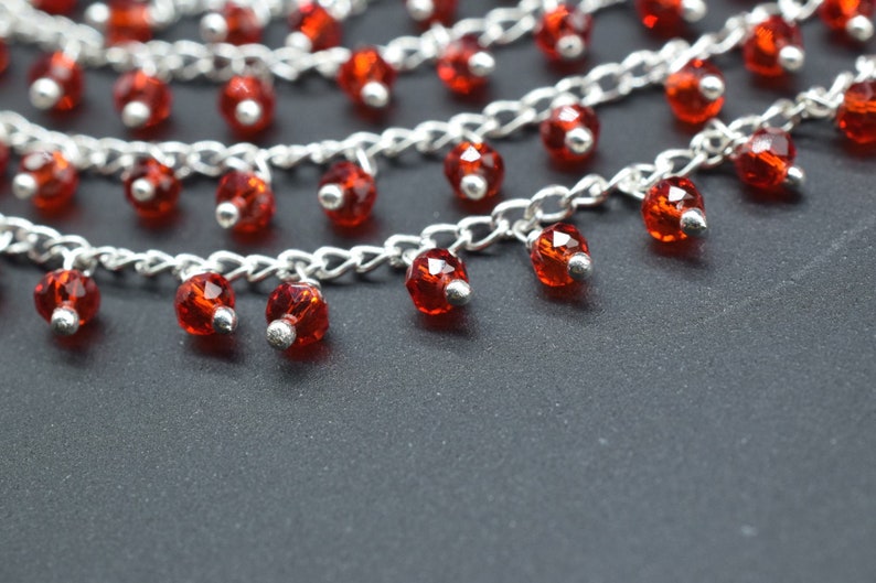 K&ouml;nnte beinhalten: Eine silberne Kettenkette mit mehreren Str&auml;ngen und roten Kristallperlen. Die Perlen h&auml;ngen an der Kette und sind facettiert.