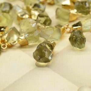 May include: A collection of raw, green gemstone pendants with gold-toned jump rings. The gemstones are irregular in shape and have a natural, rough texture.