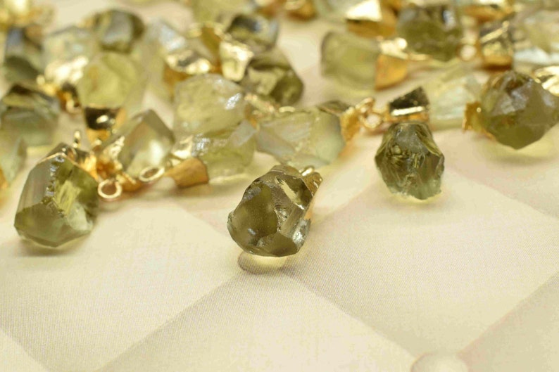 May include: A collection of raw, green gemstone pendants with gold-toned jump rings. The gemstones are irregular in shape and have a natural, rough texture.