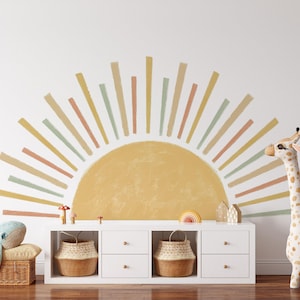 May include: A white wall with a large yellow sun with colourful rays painted on it. There is a white dresser with two drawers and two baskets underneath. A stuffed giraffe and a stuffed whale are on the floor in front of the dresser.