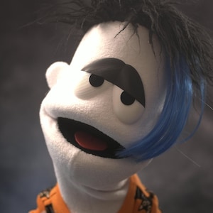 May include: A white puppet with a black eyebrow, dark hair with a blue streak, and an orange shirt. The puppet has a wide, open mouth with a red interior, round black eyes, and a textured fabric surface. The head is tilted slightly to the side.