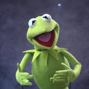 May include: A plush green Kermit the Frog puppet with large white eyes, a wide open mouth revealing a red interior, and a green collar. The puppet's arms are slightly raised, and it appears to be smiling against a dark blue background.