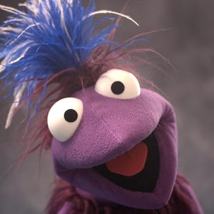May include: A purple puppet with large white eyes, black pupils, and a wide open mouth showing a red tongue. The puppet features a tuft of bright blue and white feathers on top of its head and is covered in a soft, textured purple fabric material.