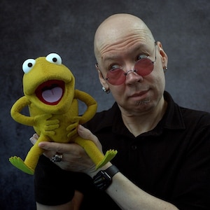 May include: A yellow frog puppet with white eyes and a red mouth is held by a person. The puppet has green feet and is smiling. The person is wearing a black shirt and round pink glasses.