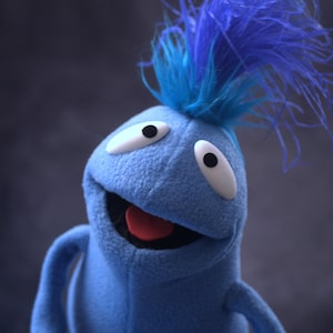 May include: A blue puppet with a smiling expression. The puppet has large white eyes with black pupils, a red tongue, and a tuft of blue feathers on top of its head. The puppet is made of a soft, plush material.