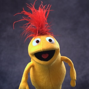 Peut inclure: Une marionnette jaune avec une crête rouge en plumes, de grands yeux blancs et une large bouche souriante. La marionnette a une bouche noire avec une langue rouge en forme de cœur et des bras jaunes.