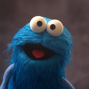 Puede incluir: Un títere de peluche azul con pelaje largo y desgreñado, dos grandes ojos esféricos blancos con pupilas negras y una boca abierta que muestra una lengua roja. El personaje tiene una expresión amplia con una frente prominente y suave textura.