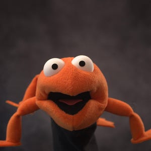May include: An orange frog puppet with large white eyes and a wide open mouth. The puppet is set against a dark gray background with red curtains on either side, creating a stage-like setting.