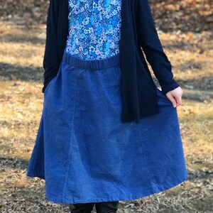 May include: A young person wearing a blue and white floral print top, a dark blue denim skirt, and a black cardigan. The person is standing in a wooded area.