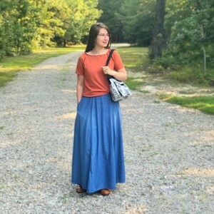 May include: A woman wearing a rust-colored t-shirt and a long denim skirt. She is carrying a white and black patterned tote bag.