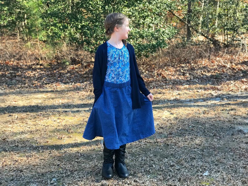 May include: A young person wearing a blue denim skirt with a floral print top and a black cardigan. The skirt has a wide waistband and pockets. The person is standing in a wooded area.