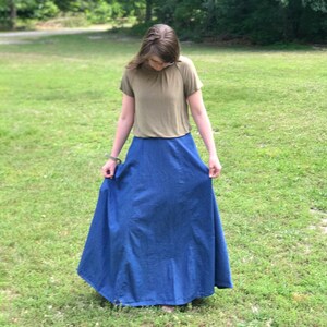 May include: A woman wearing a light brown short-sleeved shirt and a long blue denim skirt.