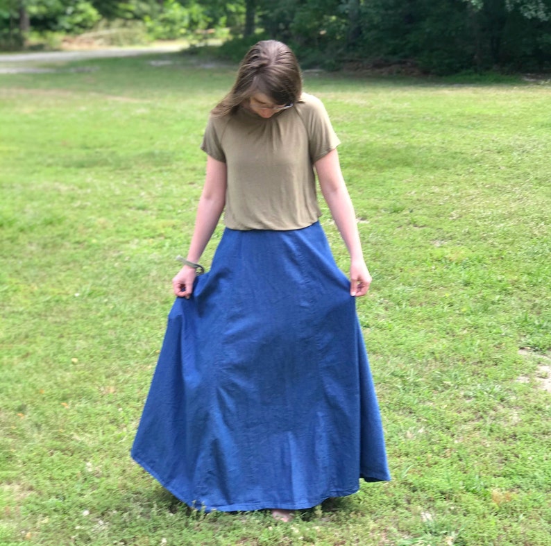 May include: A woman wearing a light brown short-sleeved shirt and a long blue denim skirt.