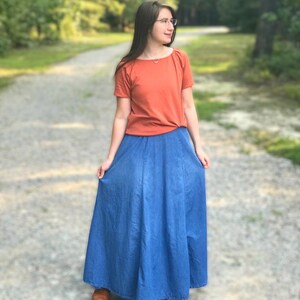May include: A woman wearing a rust-colored short-sleeved shirt and a long denim skirt. She is standing on a gravel path with trees in the background.