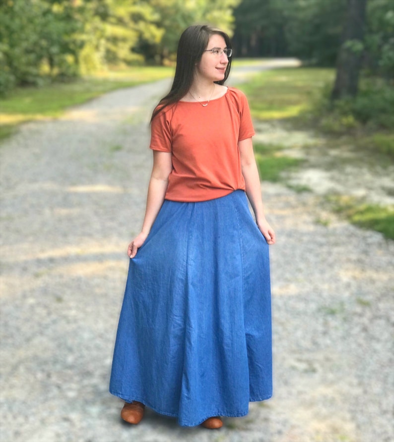 May include: A woman wearing a rust-colored short-sleeved shirt and a long denim skirt. She is standing on a gravel path with trees in the background.
