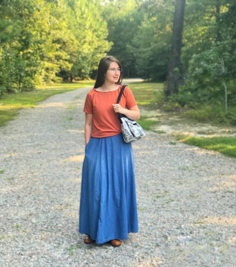 May include: A woman wearing a rust-colored t-shirt and a long denim skirt. She is carrying a white and black patterned tote bag.