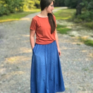 May include: A woman wearing a rust-colored short-sleeved shirt and a long denim skirt. She is standing on a gravel path with trees in the background.