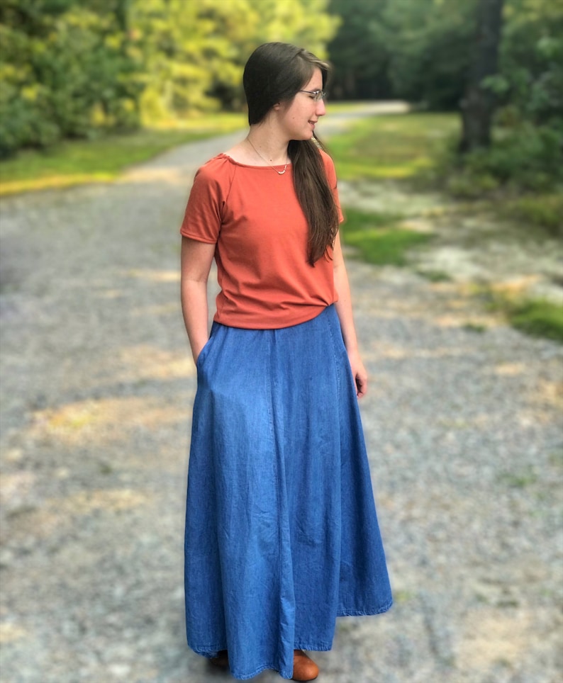May include: A woman wearing a rust-colored short-sleeved shirt and a long denim skirt. She is standing on a gravel path with trees in the background.