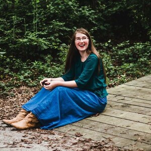 May include: A woman wearing a teal green shirt and a blue denim skirt sits on a wooden deck. She is smiling and looking to the left. The background is a green forest.