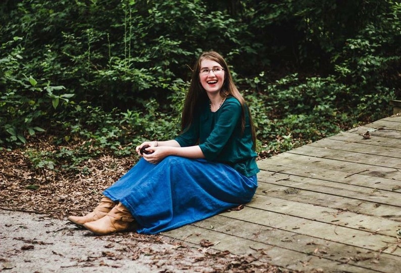 May include: A woman wearing a teal green shirt and a blue denim skirt sits on a wooden deck. She is smiling and looking to the left. The background is a green forest.