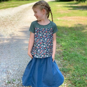 May include: A young girl wearing a green and floral print short-sleeved shirt with a blue denim skirt.