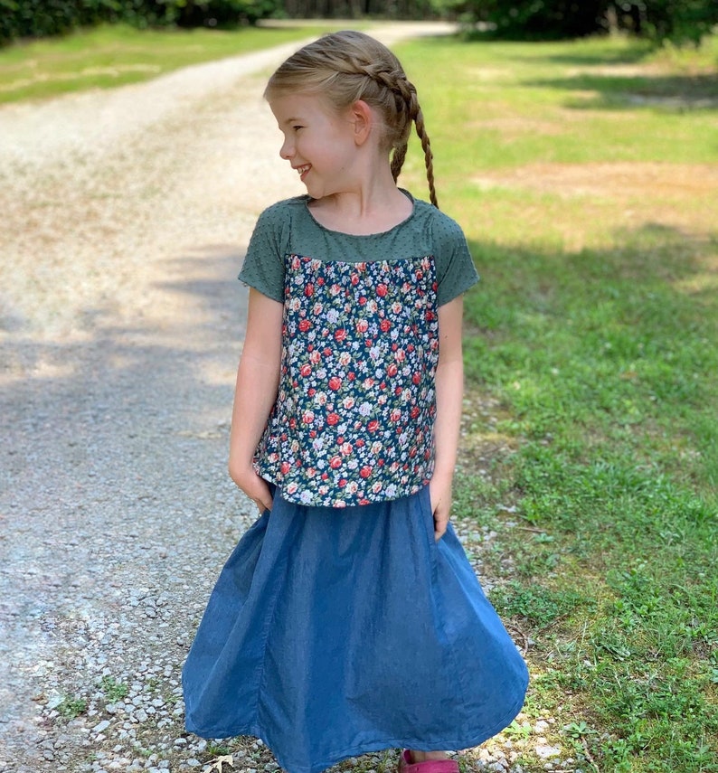 May include: A young girl wearing a green and floral print short-sleeved shirt with a blue denim skirt.