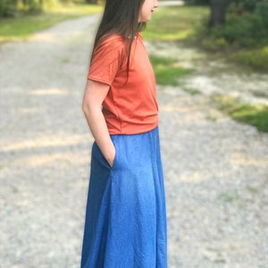 May include: A woman wearing a bright orange short-sleeved shirt and a long denim skirt with a pocket. She is standing on a gravel path with green trees in the background.