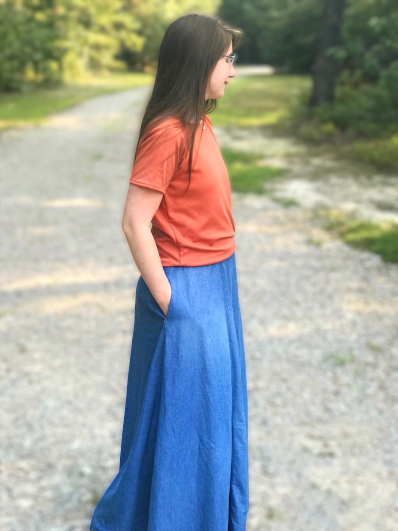 May include: A woman wearing a bright orange short-sleeved shirt and a long denim skirt with a pocket. She is standing on a gravel path with green trees in the background.