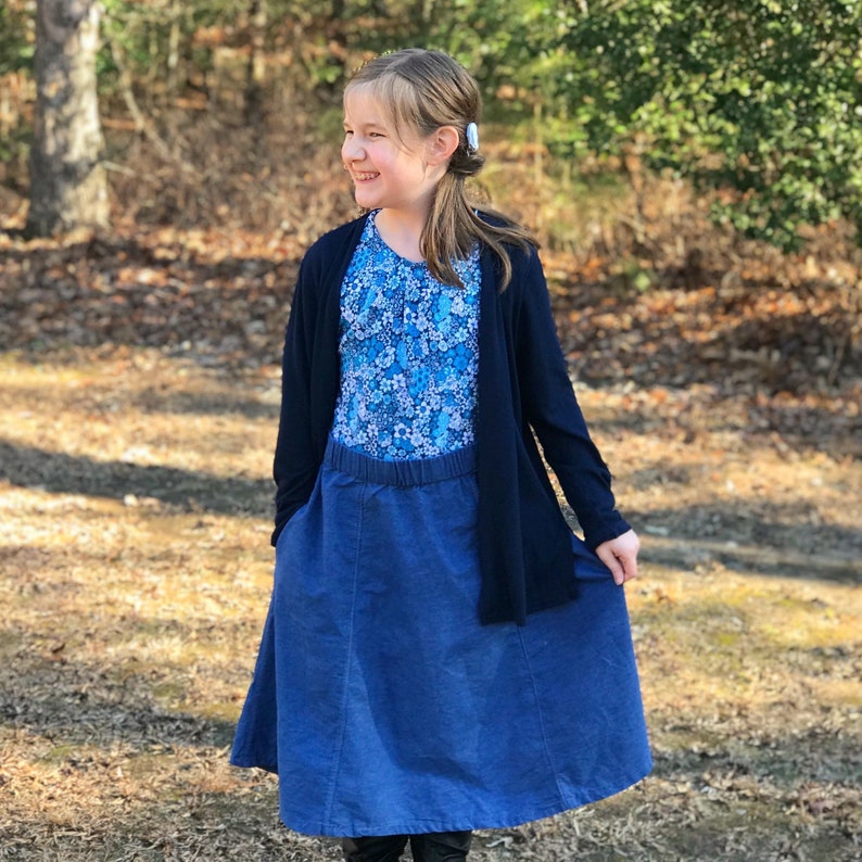 May include: A young person wearing a blue and white floral print top, a dark blue denim skirt, and a black cardigan. The person is standing in a wooded area.