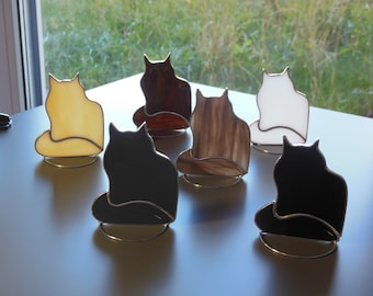 May include: A collection of six stained glass cat figurines in various colors including black, white, yellow, and brown. Each cat silhouette is outlined in metal and rests on a circular wire stand, showcasing smooth surfaces and distinct color variations.