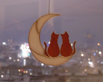 May include: A stained glass suncatcher featuring two orange cats sitting side by side on a white crescent moon. The piece is constructed with thin metal solder lines outlining the shapes of the cats and the moon, suspended by a thin wire.
