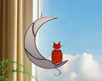 May include: A stained glass suncatcher featuring a white crescent moon and a small orange cat sitting on the curve. The cat has thin metal wire whiskers and a curved tail. The piece is suspended by a thin cord, showcasing translucent glass.