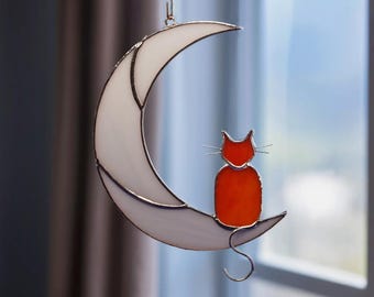 May include: A stained glass suncatcher featuring a white crescent moon and a small orange cat sitting on the lower curve. The cat has thin metal wire whiskers and a curved tail hanging down. The pieces are joined by dark metal solder lines.