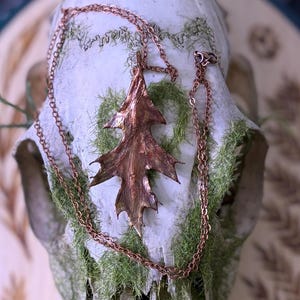 May include: A copper-colored leaf pendant necklace. The pendant is a detailed replica of a leaf, with intricate veining and a textured surface. The necklace chain is also copper-colored. The leaf pendant is displayed on a white skull.