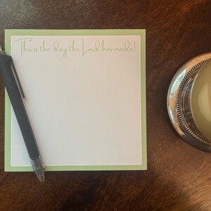May include: A notepad with a light green border and the inscription "This is the day the Lord has made!" in gold script. A black pen rests on the notepad. A glass of liquid sits on a silver coaster.