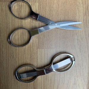 May include: Two pairs of silver-colored scissors on a wood surface. One pair has a traditional design with two loops and a pointed blade. The other pair has a more modern design with a single loop and a blunt blade.