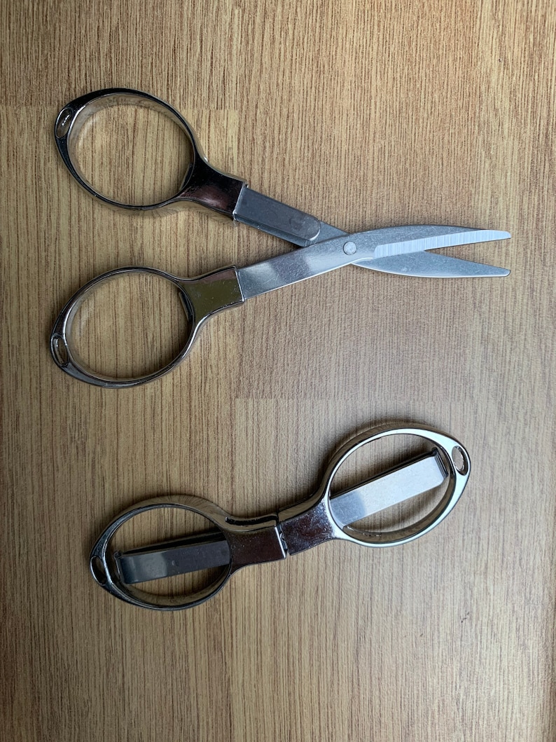May include: Two pairs of silver-colored scissors on a wood surface. One pair has a traditional design with two loops and a pointed blade. The other pair has a more modern design with a single loop and a blunt blade.