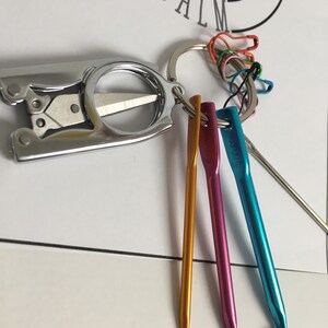 May include: A silver keychain with a small pair of scissors, a key ring, and three colorful crochet hooks. The keychain also has several safety pins attached to the key ring.