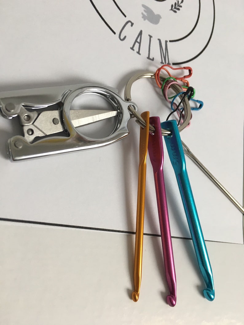May include: A silver keychain with a small pair of scissors, a key ring, and three colorful crochet hooks. The keychain also has several safety pins attached to the key ring.