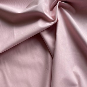 May include: Close-up of a light pink fabric with soft folds and subtle texture. The fabric appears smooth and drapes naturally, creating shadows and highlights. The overall impression is of a delicate, high-quality material, suitable for apparel or home decor.