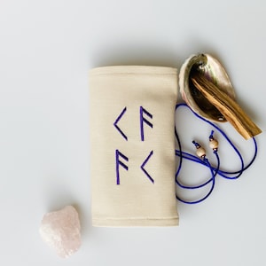 Könnte beinhalten: Ein beigefarbener Stoffbeutel mit lila gestickten Symbolen, ein Rosenquarz-Kristall, eine polierte Abalone-Schale und ein Bündel Palo-Santo-Holz. Ein blaues Band mit Holzperlen ist ebenfalls vorhanden. Die Artikel sind auf einer weißen Oberfläche angeordnet.