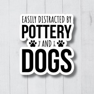 May include: White sticker with black text that reads "Easily Distracted by Pottery and Dogs" with two paw prints between the words "Pottery" and "Dogs".