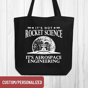May include: Black tote bag with white text that reads "IT'S NOT ROCKET SCIENCE, IT'S AEROSPACE ENGINEERING." The design includes a graphic of a rocket ship in front of a moon. The bottom of the bag has a red rectangle that says "CUSTOM/PERSONALIZED."